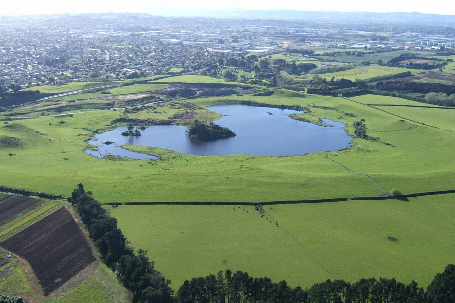 Aerial view of Crater Hill, showing the lake and wooded island within its grassy crater, surrounded by farmland and suburbs.