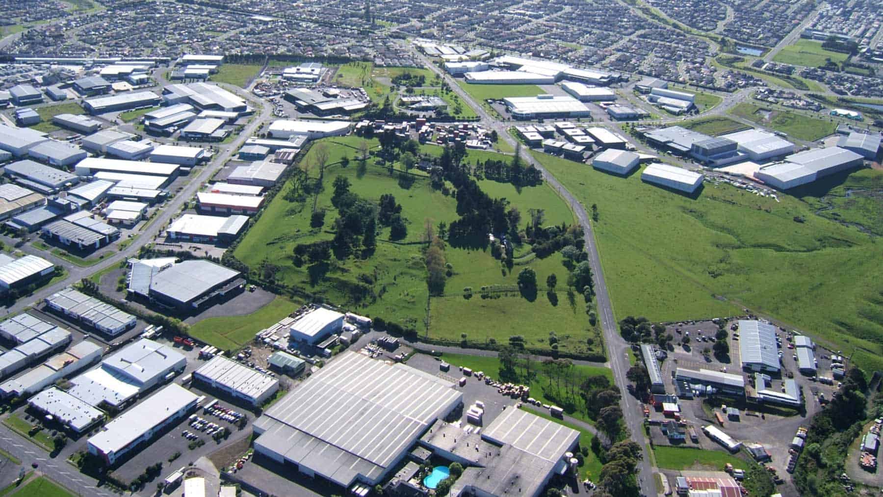 Aerial view of Hampton Park Volcano, showing its grassy volcanic landforms and trees surrounded by industrial buildings and suburbs.