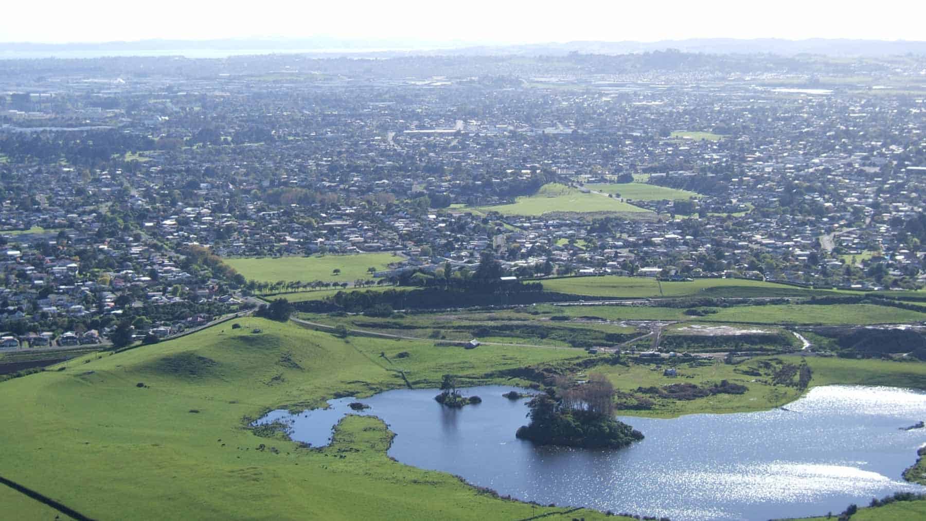 Aerial view towards Kohuora Crater and surrounding suburbs, with a lake, wooded islands and grassy farmland in the foreground.