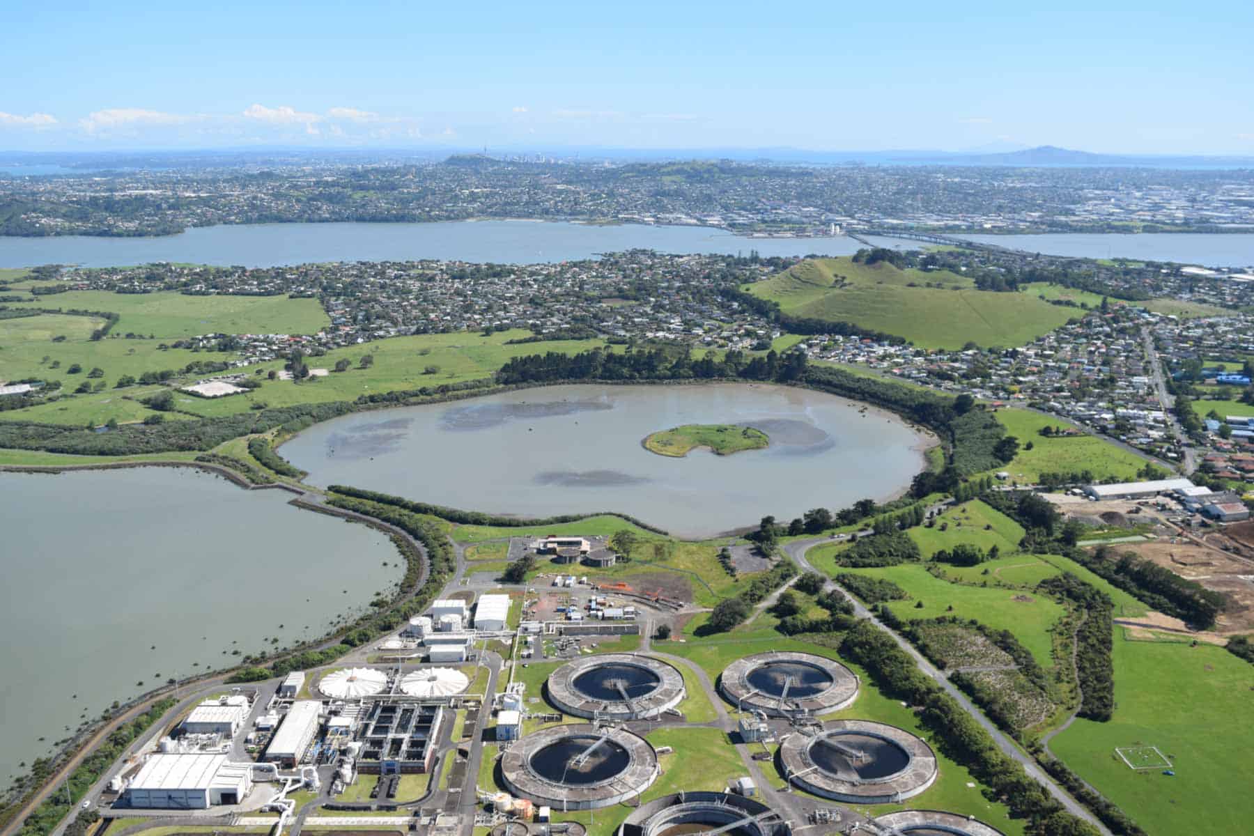 Aerial view of Māngere Lagoon and its central grassy island, with Māngere Mountain beyond and wastewater treatment tanks in the foreground.