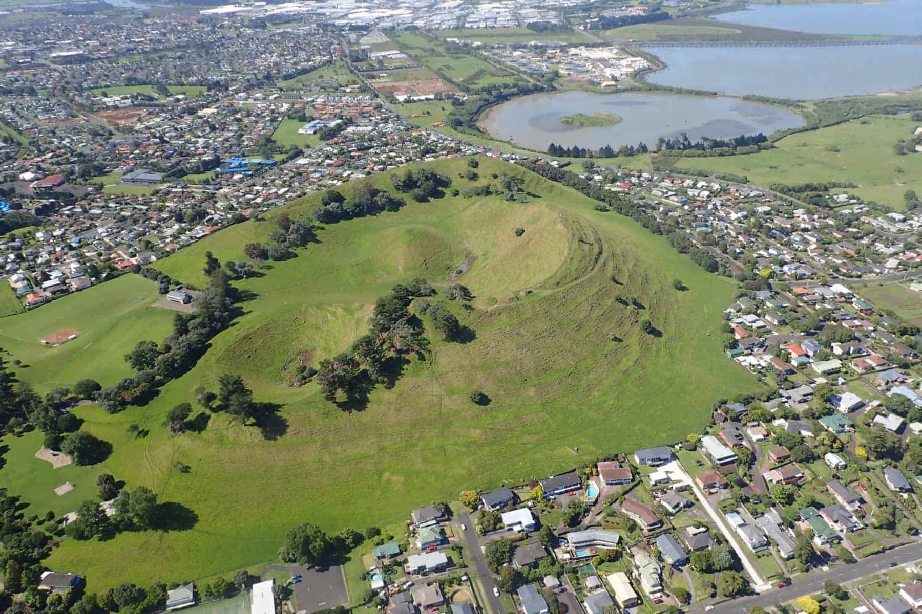 Aerial view of Māngere Mountain’s grassy volcanic crater, surrounded by homes, with Māngere Lagoon beyond.