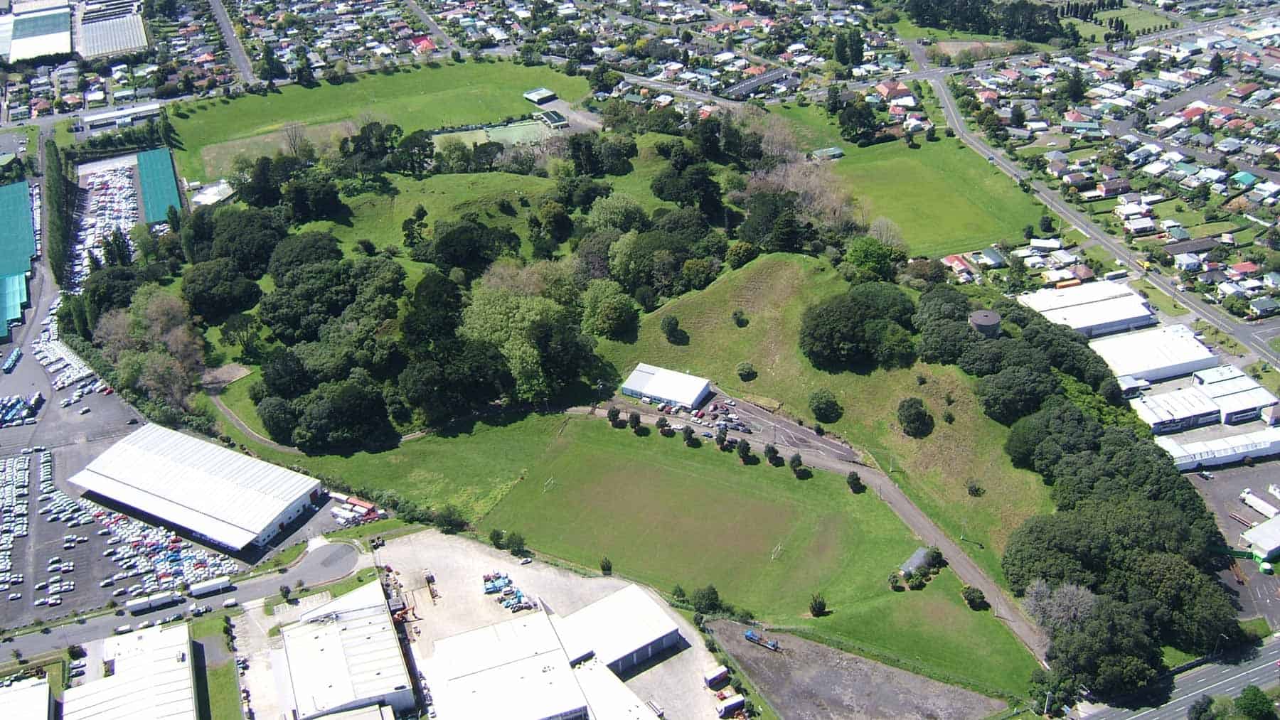 Aerial view of Ōtāhuhu / Mt Richmond, showing grassy volcanic hills, trees and sports fields surrounded by Auckland housing and industrial buildings.