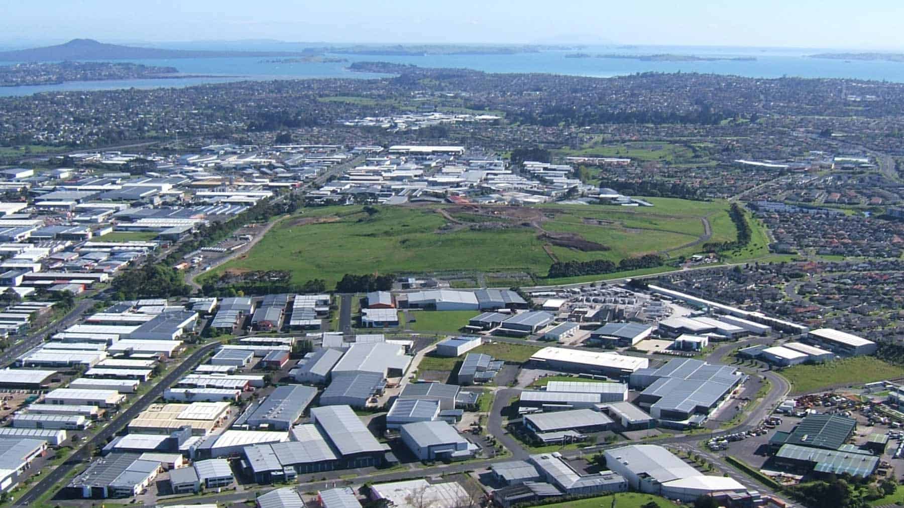 Aerial view of the grass-covered former quarry at Te Puke-o-Taramainuku / Ōtara Hill, surrounded by industrial buildings and suburbs, with the Hauraki Gulf in the distance.