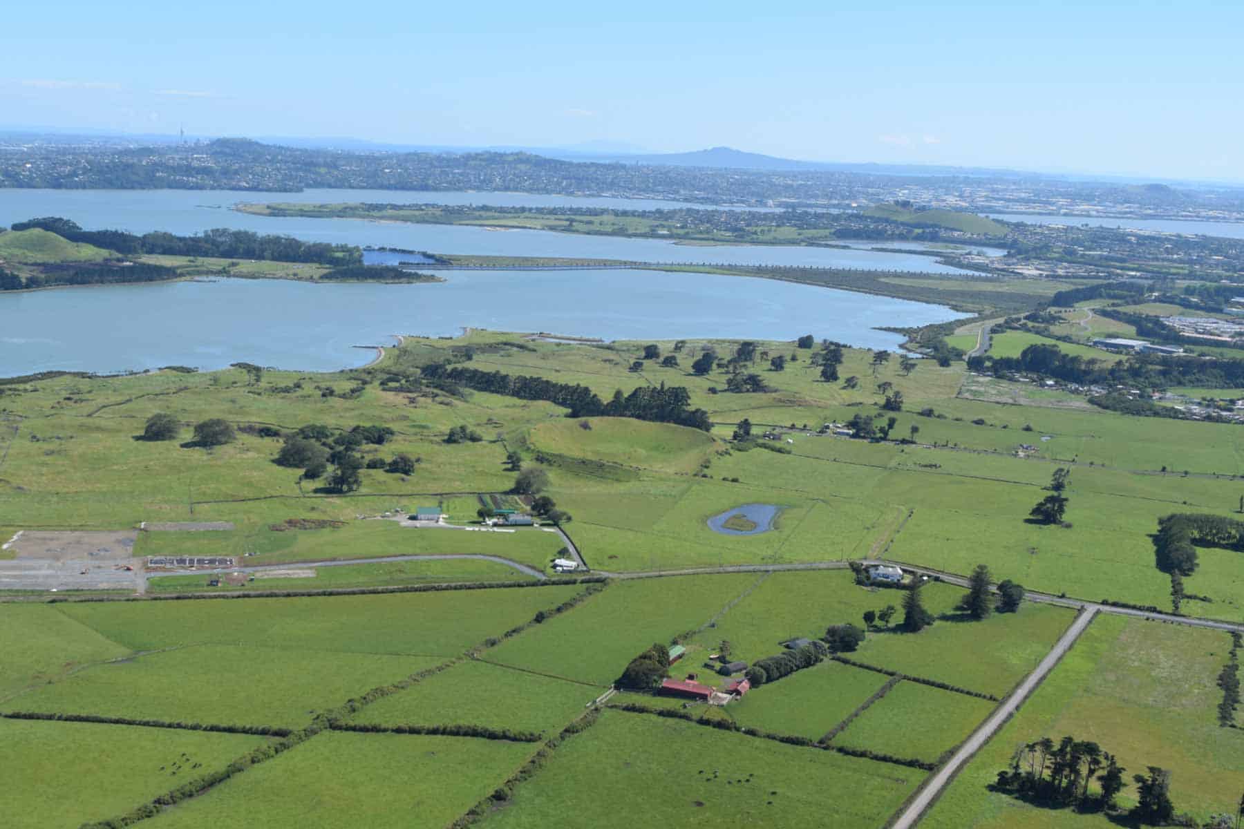 Aerial view of Ōtuataua’s grassy volcanic remnant and surrounding stone-walled fields, with the Manukau Harbour and Auckland suburbs beyond.