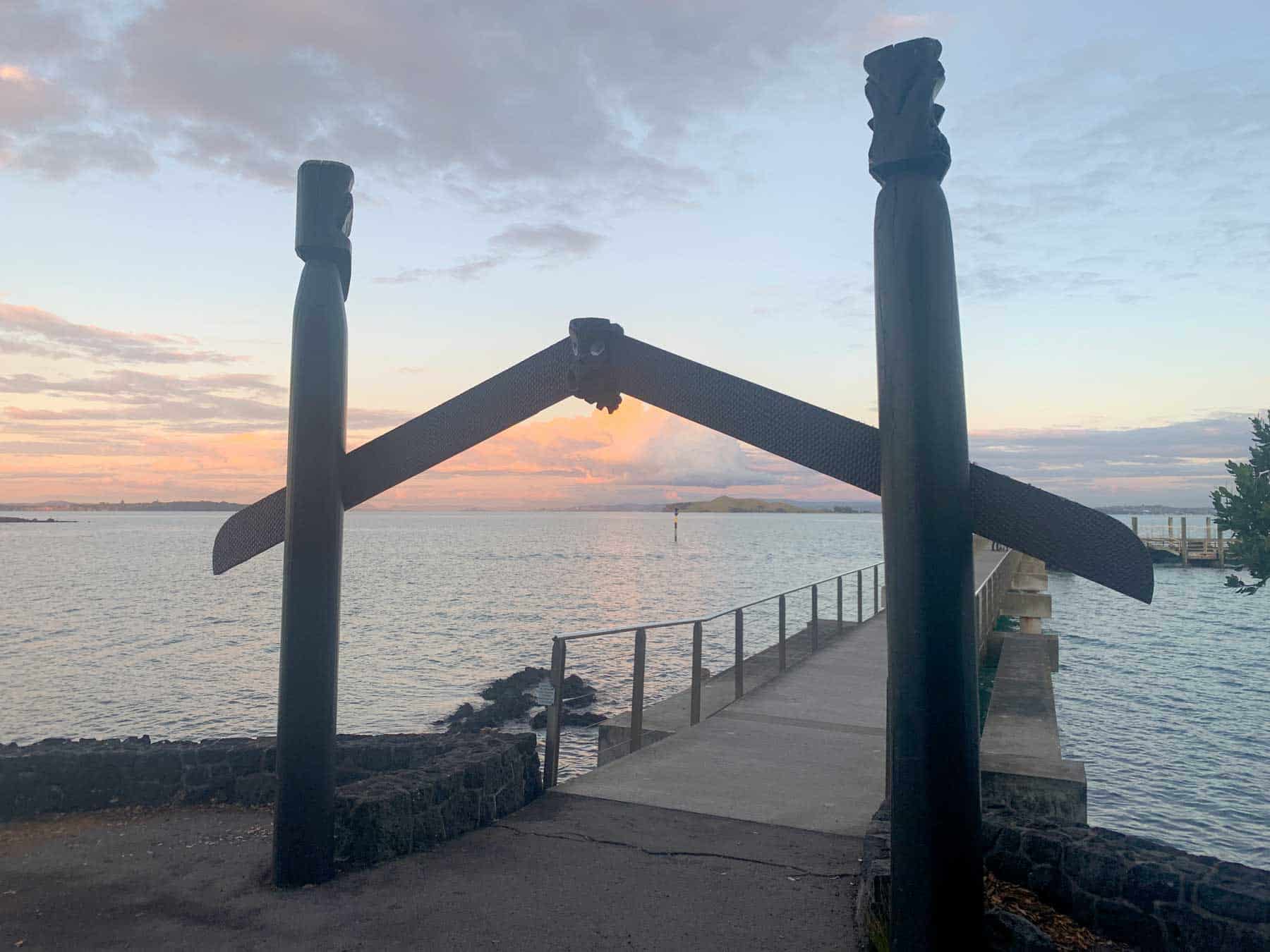 Carved Māori gateway beside the water at sunset, framing a jetty and a distant volcanic island.