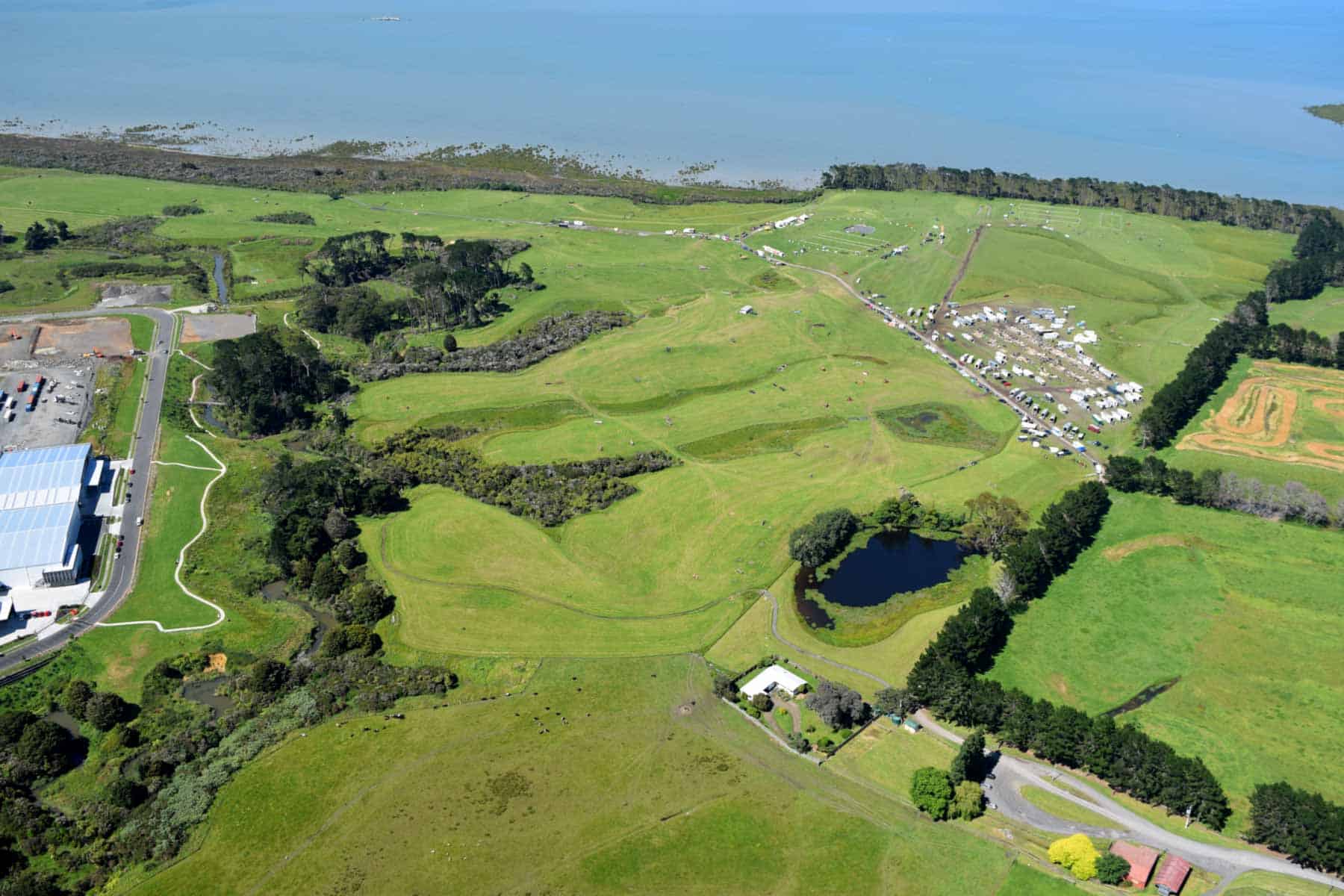 Aerial view of Puhinui Craters’ grassy tuff rings and pond, with trees, equestrian grounds and the Manukau Harbour beyond.