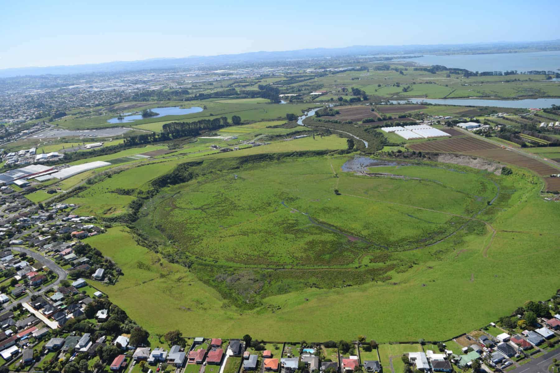 Aerial view of Pūkaki Lagoon, showing its broad grassy crater, wetland and surrounding farmland and homes, with the harbour in the distance.