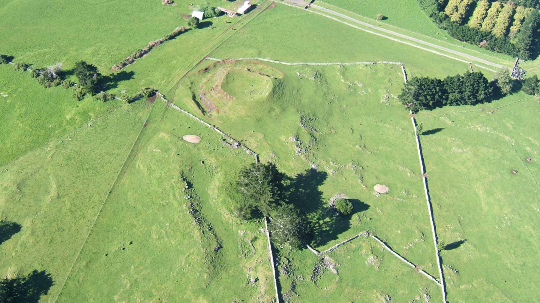 Aerial view of Pukeiti’s small grassy volcanic cone and shallow summit crater, surrounded by pasture, stone walls and scattered trees.