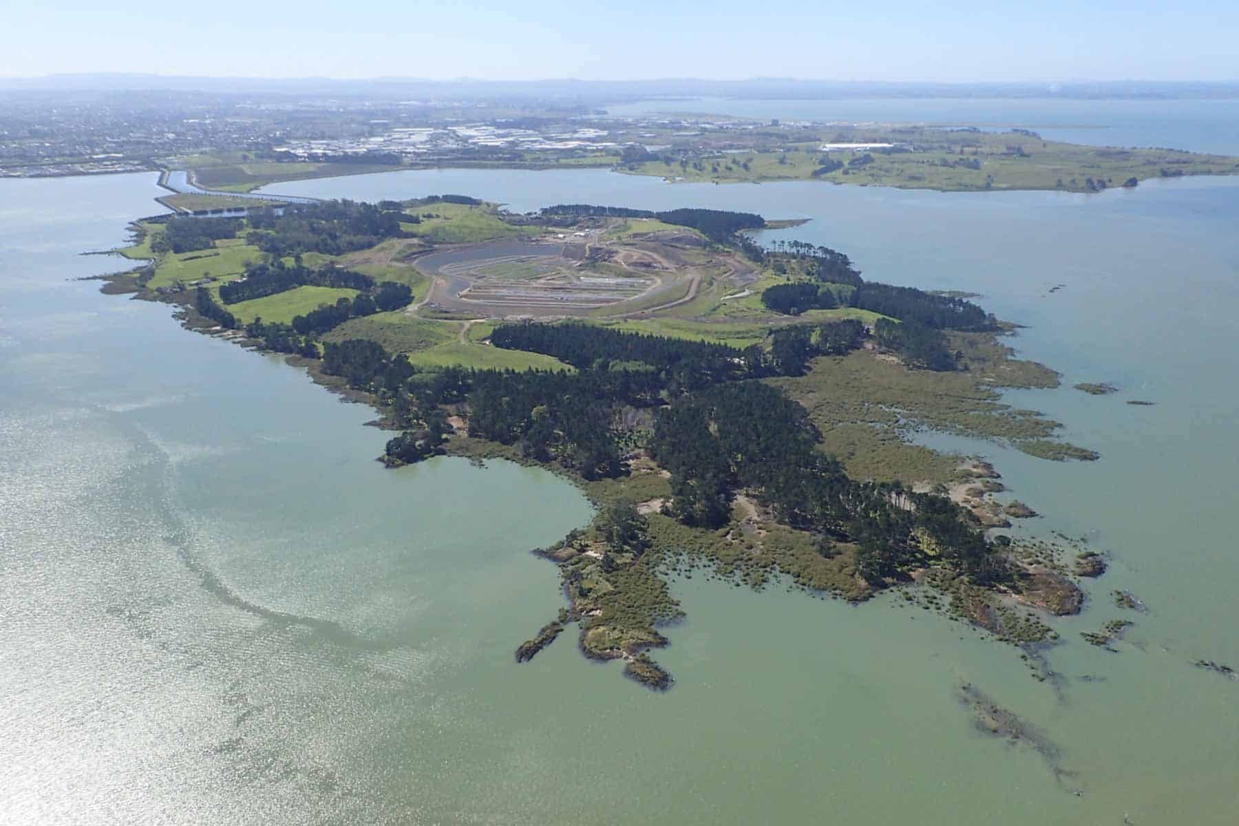 Aerial view of Puketūtū Island in the Manukau Harbour, showing its grassy slopes, trees, rocky shoreline and terraced former quarry.