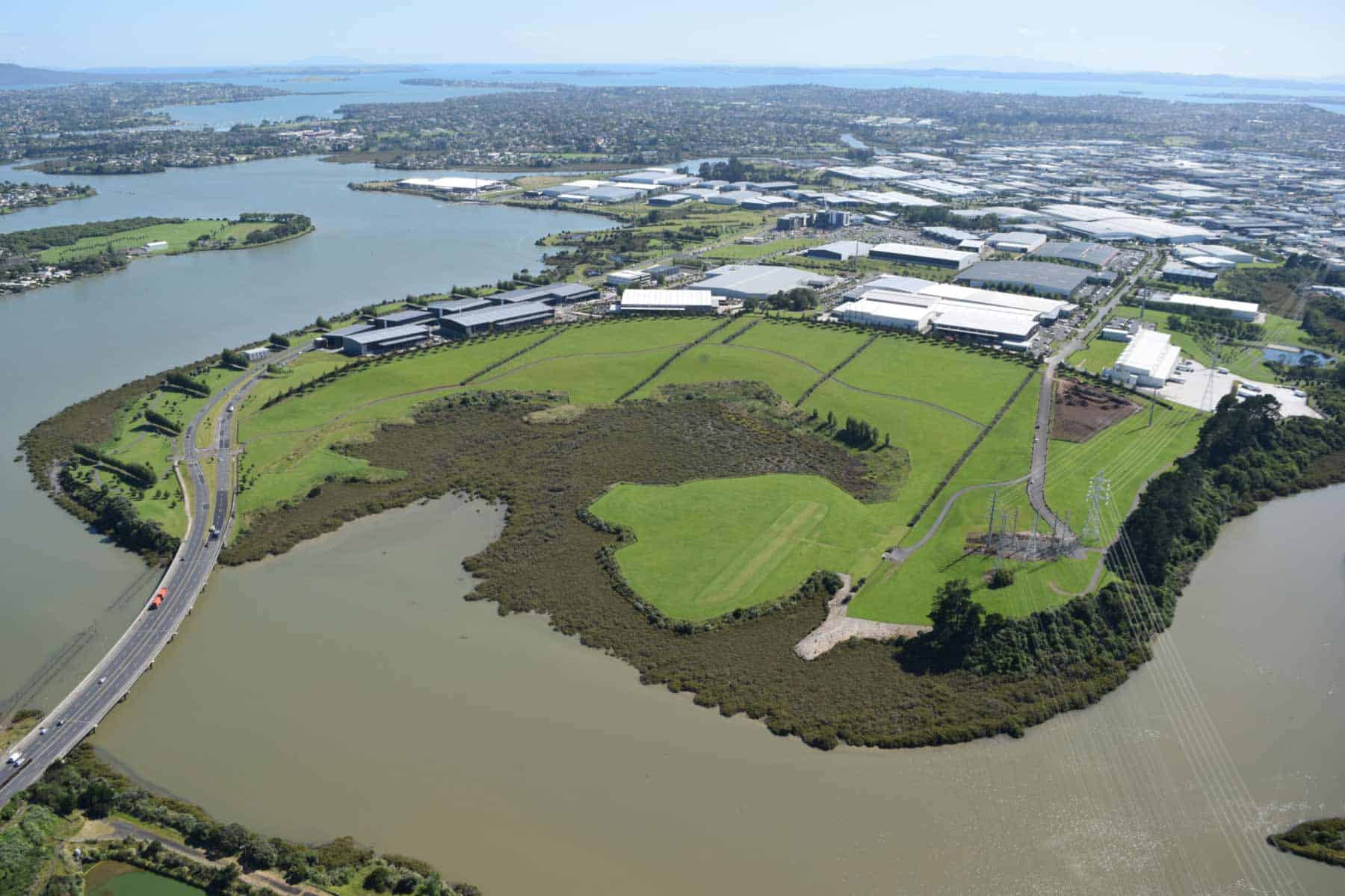 Aerial view of Pukewairiki / Highbrook Park, showing the mangrove-fringed crater, grassy parkland and surrounding waterways beside industrial buildings.