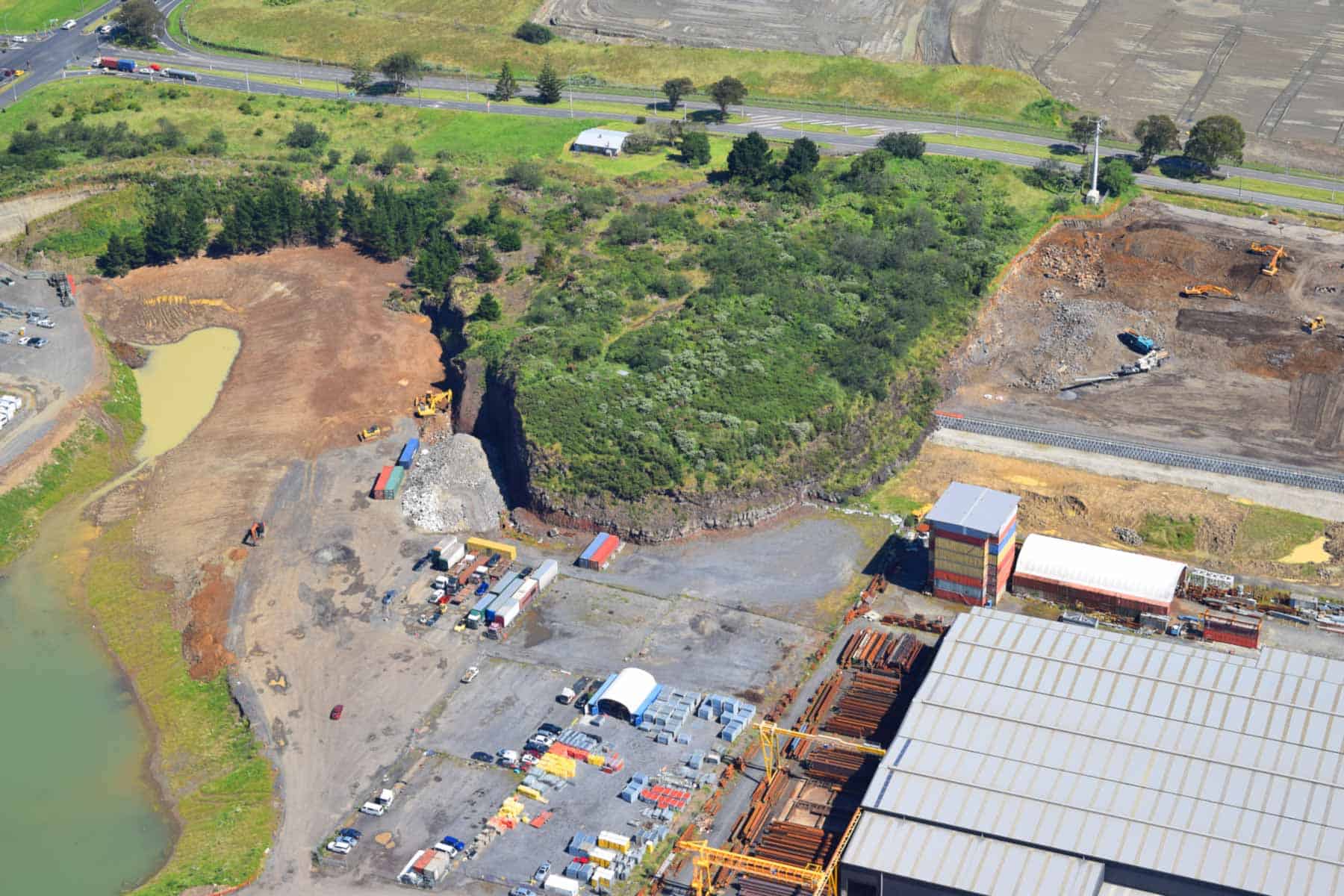 Aerial view of the vegetated remnant of Wiri Mountain, surrounded by quarry excavations, ponds and industrial buildings.