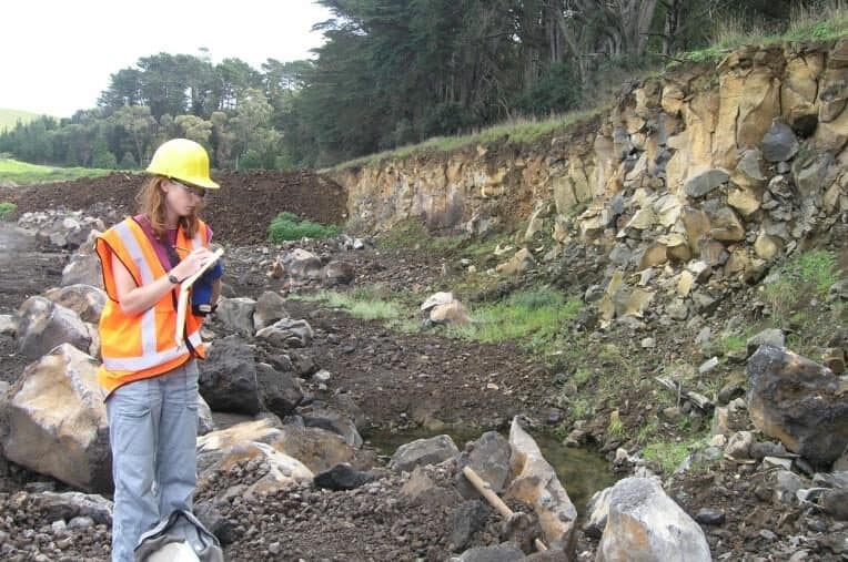 Lucy McGee, in a hard hat and high-visibility vest, taking notes in the field beside an exposed rock face
