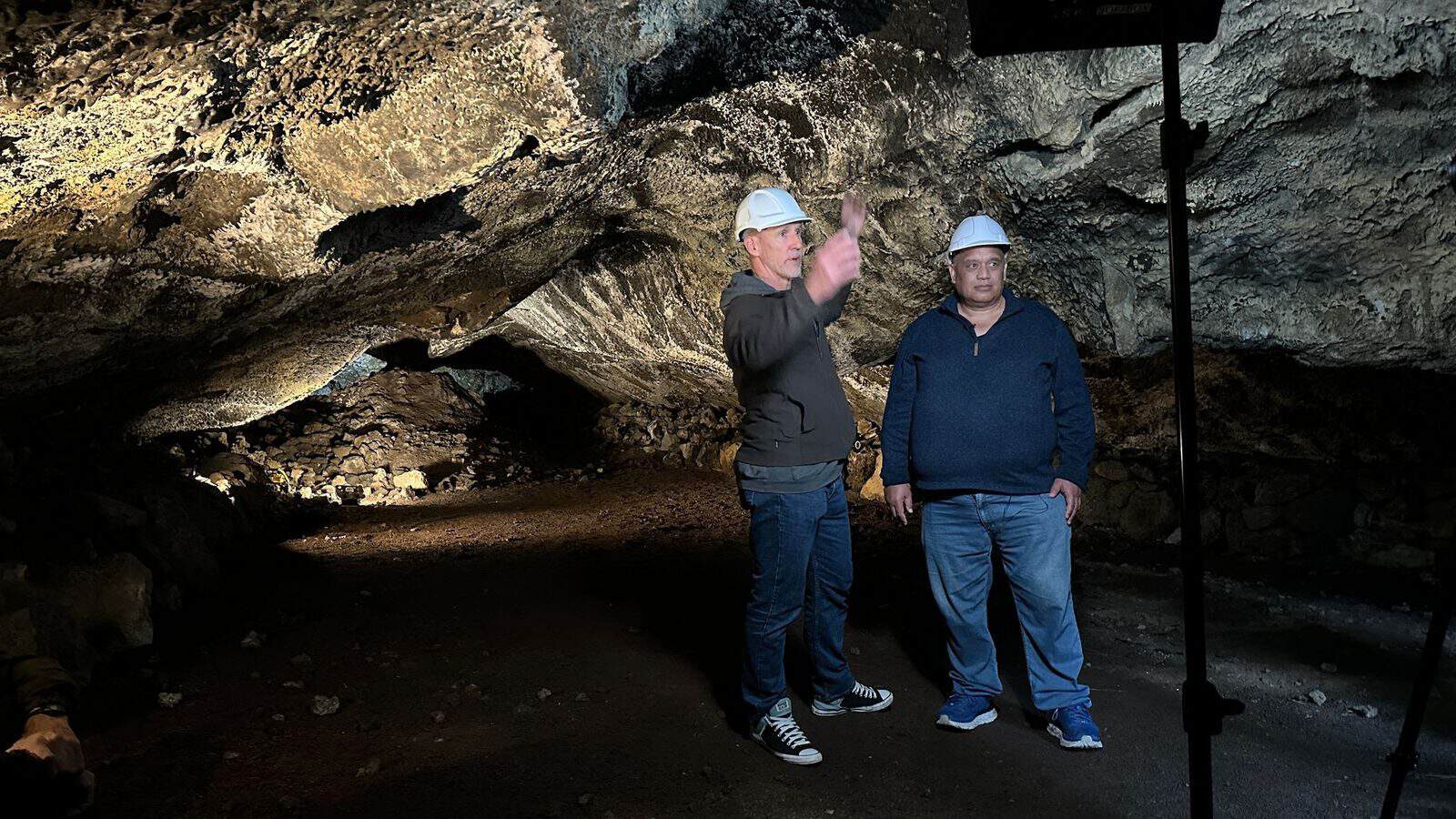 Two men in hard hats inside a lava cave, one pointing up at its rough rock ceiling